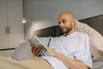 Attractive man passionately engrossed in a book he is reading while lying on a bed in a cozy bedroom, surrounded by light coming through the window, creating an atmosphere of calm and concentration