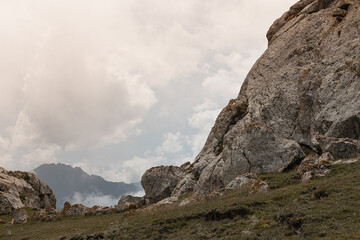 Big grey stony rock with lichen, cracks and  dim green meadow with dense white clouds in sky and silhouette dark ridge in mountains in dull frowning summer day in Dagestan. Trip in Caucasian mountain.
