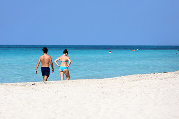 Fototapeta na wymiar Couple in swimwear walking by white sand beach against the ocean waves. Man and woman together, romantic leisure on tropical coast