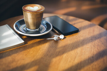 Heart shape of latte coffee in old style glass and mobile phone, note book, wireless headphones on wooden table with vintage sofa and sunlight