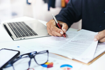 Close-up view, successful businessman, manager or freelancer holding pen to review document on desk at office, using laptop to meeting and work online