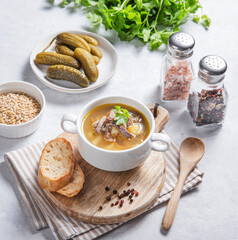 Pickle soup with pearl barley, pickles and meat in a bowl on a wooden board with bread and fresh herbs.