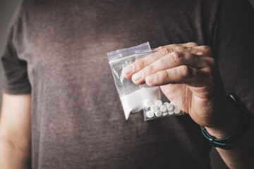 Man's hand holds two transparent plastic packets with cocaine powder and white pills lsd, drug dealer selling drugs. Drug abuse and addiction concept - obrazy, fototapety, plakaty