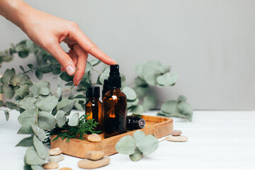 Female hand pushes on amber spray bottle with facial cosmetics on a tray with a branch of eucalyptus on the gray wooden table, facial cleaner, serum. Front view