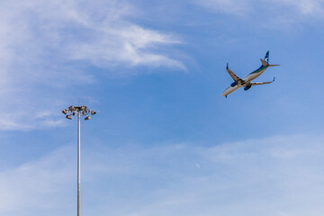 The plane flies next to a pole with searchlights against the blue sky