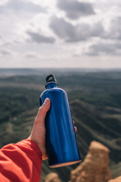 Vertical image of an anonymous hiker's hand holding a blue metal water bottle on a rocky cliff.r