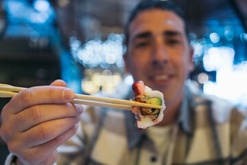 Close-up image of a young man holding a freshly made piece of sushi with wooden chopsticks.