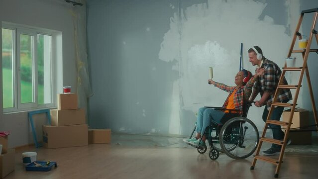A white man wheeling an African American woman in a wheelchair into the room. A young couple in big headphones enjoys music, sings using a roller as a microphone and laughs.