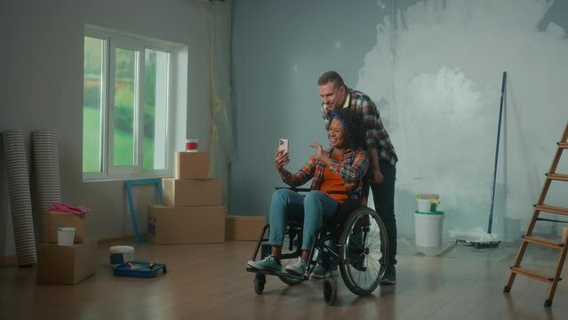 A white man and an African American disabled woman in a wheelchair are talking on video call using a mobile phone. A young couple communicates and shares plans for repair the room.