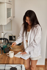 Young happy brunette woman making hot tasty beverage coffee, sweet breakfast, reading book - 576633432