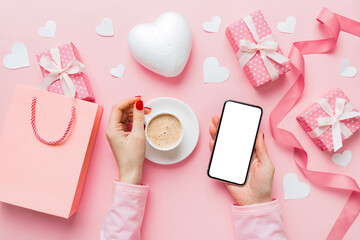 Female hands holding smart phone with coffee for Valentine day, gift box and envelope, hearts