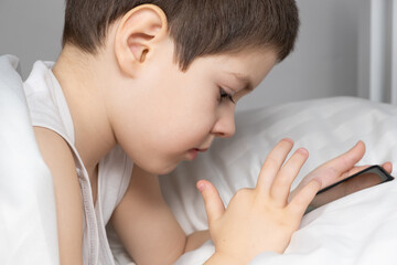 A 5 year old boy plays games on his phone while lying in bed. Gadgets in bed before bedtime in a child