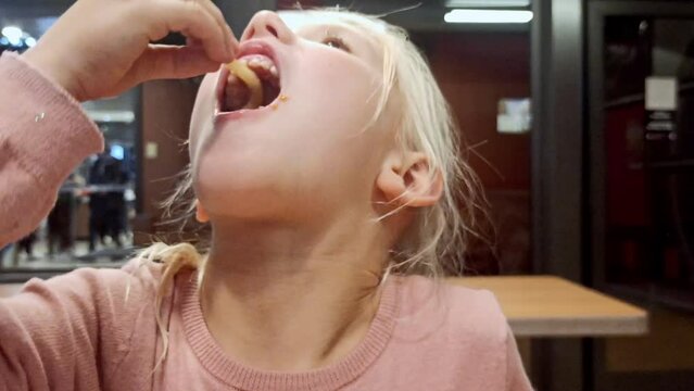 Blonde pigtail girl enjoys biting a burger and french fries in restaurant. Close-up