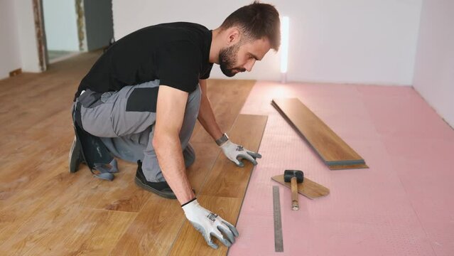 Using the hammer. A man in grey uniform lays a laminate in a room.