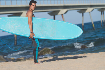 Hispanic surfer boy standing on the beach in neoprene holding his blue surfboard