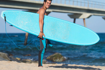 Hispanic surfer boy standing on the beach in neoprene holding his blue surfboard