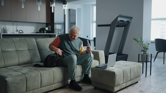 Senior Man Stroking Black Cat On Sofa And Doing Exercise With Dumbbell, Home Fitness And Healthy Lifestyle