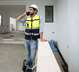 Engineer, bearded man wears safety helmet communicating housing development project at construction site using smartphone. Contractor manager examining residential building estate infrastructure.