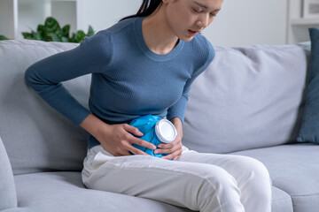 Asian woman using hot water bottle to relieve abdominal pain on sofa at home. Sick female having a...