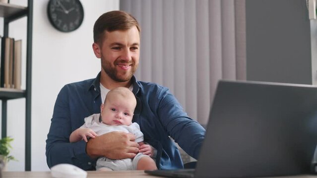 Sitting by table and using laptop. Father with his little baby is at home.