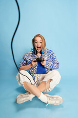 Happy excited young girl emotionally playing video games, sitting over blue studio background. Leisure time activity. Concept of youth, emotions, lifestyle, facial expression, hobby, fun