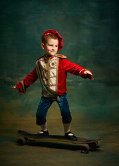 Smiling little boy dressed up as medieval little prince and pageboy skateboarding over dark vintage...