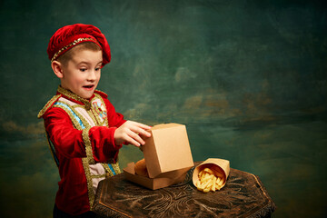 Happy cheerful little boy wearing costume of medieval page boy and prince eating hamburger with french fries over dark green background.
