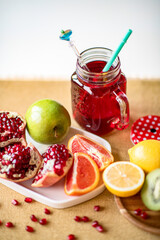 Fresh pomegranate juice decorated with staw in glass mug and sliced colorful fruits: orange, green apple, yellow lemon, kiwi and chopped pomegranate on white plate. Healthy eating and drinking