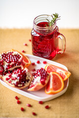 Fresh pomegranate juice decorated with green rosemary in glass mug, sliced red orange and chopped pomegranate on white plate. Healthy eating and drinking