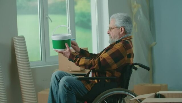 An elderly disabled man in a wheelchair takes a bucket of paint, roller and plans to paint the walls. A pensioner plans repairs and wall decoration. Handicapped person.
