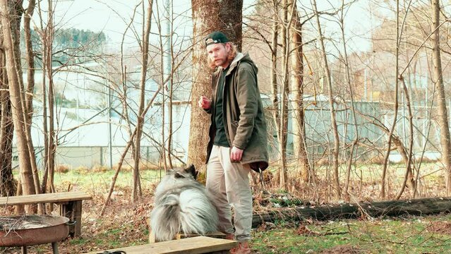 This young man commands his fluffy gray dog to sit down. The wolfspitz seems to be very disciplined. After completing the exercise and placing the dog, his owner looks towards the camera