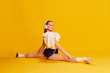 Hobby. Beautiful young girl in headphones and sports wear sitting on twine, training, stretching over yellow studio background. Concept of beauty, sportive lifestyle, hobby, emotions. Ad