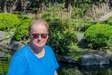 Naklejka na ściany i meble Portrait of handsome dutch senior adult man wearing sunglasses and blue blouse, pond, green bushes and trees in blurred background, enjoying sunny day in Japanese garden, looking at camera