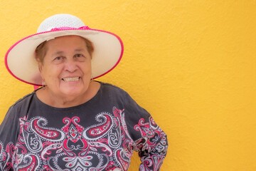 Waist-up portrait of smiling senior woman against yellow background, looking at camera, grandmother wearing hat and enjoying sunny day. Concept of enjoying life in old age. space for text