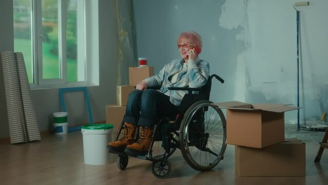 An elderly disabled woman moves in a wheelchair and talks on a mobile phone. Granny inspects a bucket of paint and pulls a roller out of a cardboard box. Room with window, ladder, wallpaper rolls.