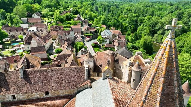 Small, medieval village situated next to a river, flowing through a rich forest in the heart of France. Droneshot flies over the village, showing medieval brick buildings, and walls overgrown by ivy.