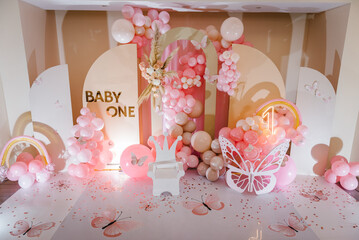 Birthday party for 1 year old girl on a background photo wall. Arch decorated pink balloons, rainbow, text baby one, flowers, paper decor butterfly, and wooden white chair. Children's photo zone. Top.