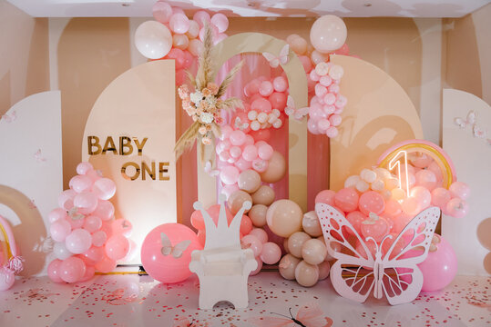 Arch decorated pink balloons, rainbow, text baby one, flowers, paper decor butterfly, and wooden white chair. Birthday party for 1 year old girl on a background photo wall. Children's photo zone.
