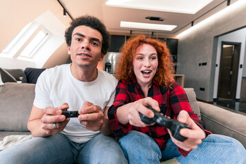 An interracial couple sits in the living room playing games, it looks like she's winning and he's stressing out