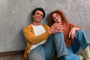 Multiracial friends sit on the floor, leaning against the wall, dressed in casual clothing, smiling and laughing