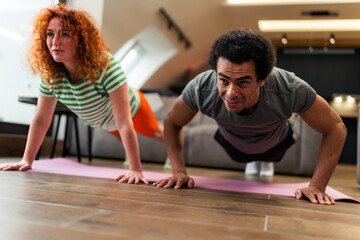 An interracial couple doing push ups together in the living room, is dressed casually and they like...
