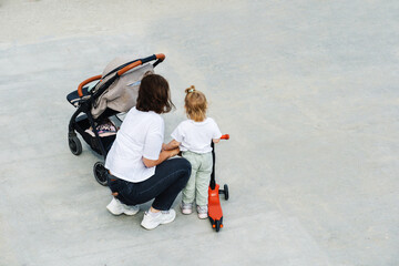 Mother and little daughter on a walk. The child on the scooter next to the parent receives...
