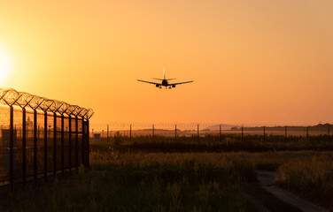 Naklejka na ściany i meble Airport. Barbed wire fence. Beautiful orange sunset. Landing plane silhouette.