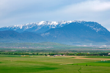 Fototapeta na wymiar Beautiful aerial view of the snowcapped Piatra Craiului mountains (Brasov, Romania) from Rasnov in springtime. Idyllic alpine mountain scenery with large green valley, villages and high mountains