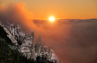 Dramatic evening winter mountains landscape. Erroded peaks or stone cliffs rise high in the foreground, background with fluffy orange clouds and sunset sun. Aerial view of snow covered mountains