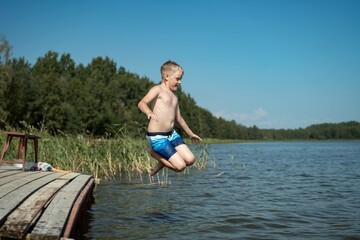 Fototapeta premium cute caucasian boy jumping from wooden pier diving into lake in countryside