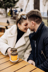 Happy young couple in love in outerwear sitting at a table outside and drinking coffee. Relaxing in the open air