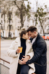 Happy young couple in love in outerwear walking on the street and drinking coffee. Recreation in the open air. A young married couple