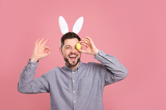 Happy man in bunny ears headband holding painted Easter egg on pink background, space for text
