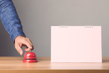 Hand ringing the red call bell and Empty blank page of paper desk calendar on the top of wooden table with grey color space wall background.